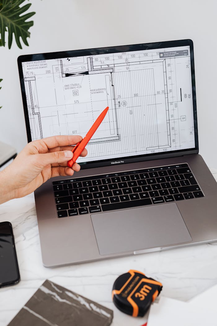 hero-img-02 A hand using a red pen points at a floor plan displayed on a laptop, showcasing architectural design.